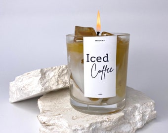 May include: A clear glass candle jar with a white label that reads "MILKYS Iced Coffee 180G". The candle is lit and the wax is a light brown colour. The candle is sitting on two white rocks.