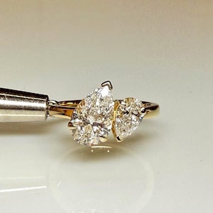 Può includere: Un anello in oro giallo con due diamanti taglio a goccia posizionati fianco a fianco. Il diamante più grande si trova a sinistra, con un diamante taglio a goccia più piccolo accanto. La fascia è sottile e lucida, con una finitura classica.