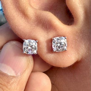 May include: Close-up of two square-cut, clear gemstone stud earrings. The earrings are set in silver metal and are worn in the ear. The gemstones have a brilliant cut, reflecting light.