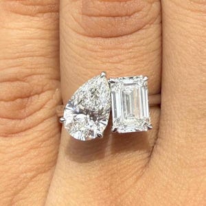 Könnte beinhalten: Nahaufnahme eines Verlobungsrings mit zwei Steinen. Er zeigt einen birnenförmigen Diamanten und einen Smaragd-Schliff-Diamanten, die auf einem silbernen Band gefasst sind. Die Diamanten funkeln und unterstreichen ihre Klarheit. Der Ring wird an einem Finger gezeigt.