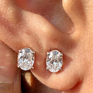 Puede incluir: Primer plano de dos pendientes de diamantes con corte ovalado. Los diamantes están engastados en una montura de plata u oro blanco. Los pendientes se llevan en una oreja, centrándose en las brillantes piedras preciosas.