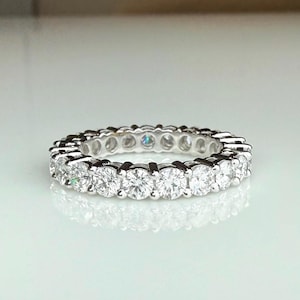 Puede incluir: Un anillo de tipo alianza en tono plateado con una hilera continua de piedras redondas transparentes de talla brillante engastadas en garras. La banda metálica tiene un acabado pulido y las piedras rodean toda la circunferencia del anillo.