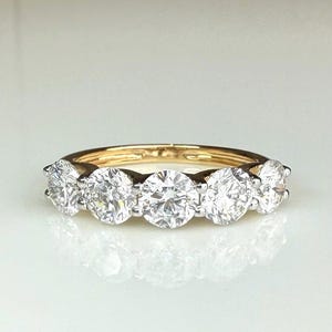May include: A five-stone ring featuring round brilliant-cut clear gemstones set in a polished yellow gold band. The stones are arranged in a horizontal row, with the centre stone being the largest, flanked by two smaller stones on each side.