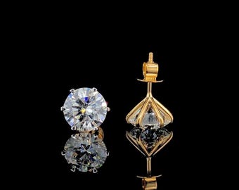 May include: Diamond stud earrings with a gold setting. The earrings feature a round-cut diamond, reflecting light. The gold setting has a distinctive geometric design. The earrings are presented on a reflective black surface.
