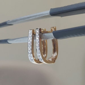 Könnte beinhalten: Ein Paar goldfarbene Creolen mit rechteckiger Form. Eine Seite jedes Ohrrings ist mit kleinen, klaren Edelsteinen besetzt. Die Ohrringe werden vor einem neutralen Hintergrund präsentiert.