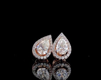 May include: A pair of pear-shaped diamond stud earrings. Each earring features a large, clear pear-cut diamond surrounded by a halo of smaller diamonds. The earrings are set in a rose gold setting, and are displayed against a black background.