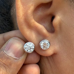 Könnte beinhalten: Ein Paar runde, klare Diamant-Ohrstecker, gefasst in einer silbernen Lünette. Ein Ohrring befindet sich im Ohr, der andere wird von einer Hand gehalten. Die Ohrringe sind ein klassisches Schmuckstück.