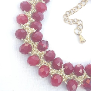 Op de afbeelding: Close-up van een bordeauxrode kralenarmband met een goudkleurige ketting en sluiting. De kralen zijn facetgeslepen en gerangschikt in een clusterpatroon, verbonden door goudkleurig draad.