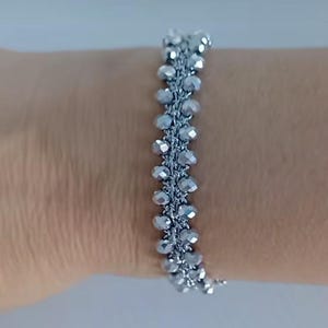 Op de afbeelding: Een zilveren armband met een geweven ontwerp. De kralen zijn facetgeslepen en hebben een subtiele glans.