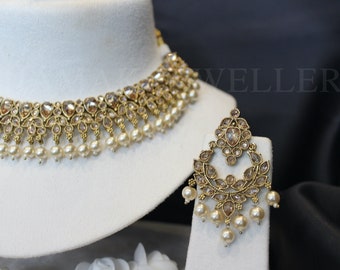 Pode incluir: Um colar choker dourado com pedras transparentes e detalhes de pérolas, combinado com brincos combinando. O colar tem um design em camadas com uma fileira de pérolas na parte inferior.
