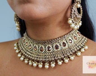 Pode incluir: Um colar choker dourado com um design de lágrima e detalhes de pérolas. O colar é adornado com cristais transparentes e marrons. O colar é combinado com brincos combinando.