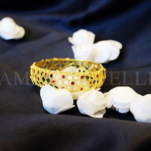 May include: Gold bangle bracelet with a delicate filigree design and red gemstones. The bracelet is surrounded by white flower petals.