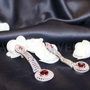 Op de afbeelding: Een paar zilveren oorhangers met rode edelstenen en heldere strass steentjes. De oorhangers liggen op een zwarte achtergrond met witte bloemen.