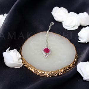 May include: A silver nose ring with a red gemstone and clear rhinestones. The ring is on a white marble-like surface with a gold rim.