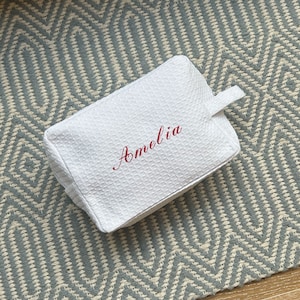 Puede incluir: Bolsa de cremallera blanca con textura de gofre con el nombre "Amelia" bordado en rojo en la parte delantera.