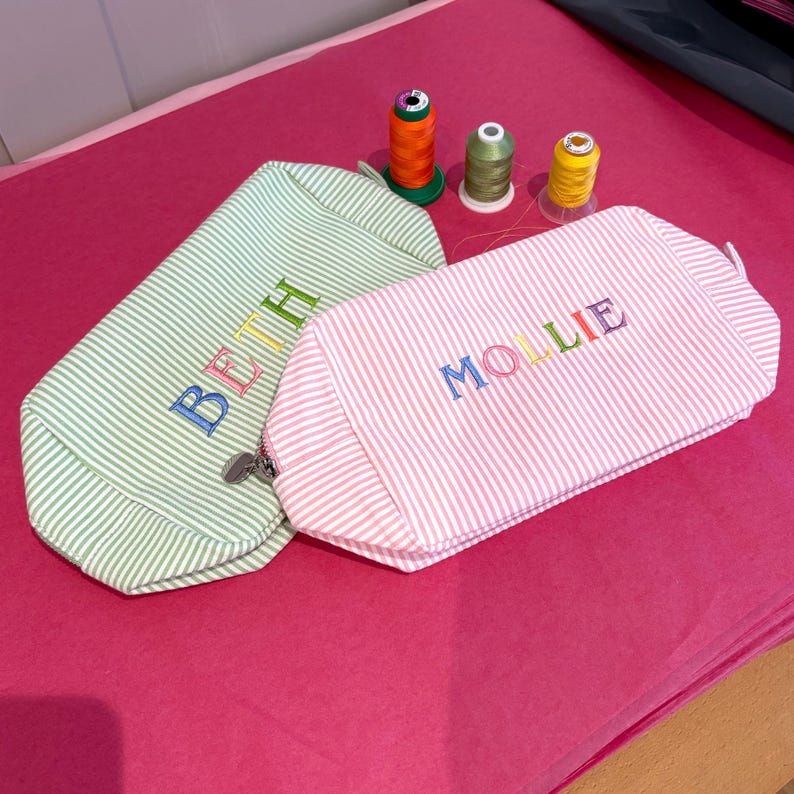 May include: Two striped fabric cosmetic bags with the names "Beth" and "Mollie" embroidered in colorful thread. The bags are on a pink surface with spools of thread.