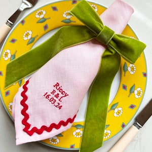 Pode incluir: Um guardanapo de linho rosa com uma borda ondulada vermelha e bordado personalizado que diz "Riccy B&L 16.03.24". O guardanapo está amarrado com um laço de veludo verde.