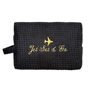 May include: Black travel pouch with a textured waffle weave pattern. The pouch features a gold embroidered airplane and the words "Jet Set & Go". It has a small handle on the side and a zipper closure.