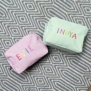 May include: Two striped fabric cosmetic bags with personalized names. The bag on the left is pink with the name "EVIE" embroidered in rainbow colors. The bag on the right is green with the name "INDYA" embroidered in rainbow colors.
