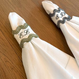 May include: Two white linen napkins, each held by a decorative napkin ring. One ring features the embroidered initials "E.L" in black thread, framed by a green and white wavy border. The other ring has the initials "S.J.G" in black, with a black and white wavy border.