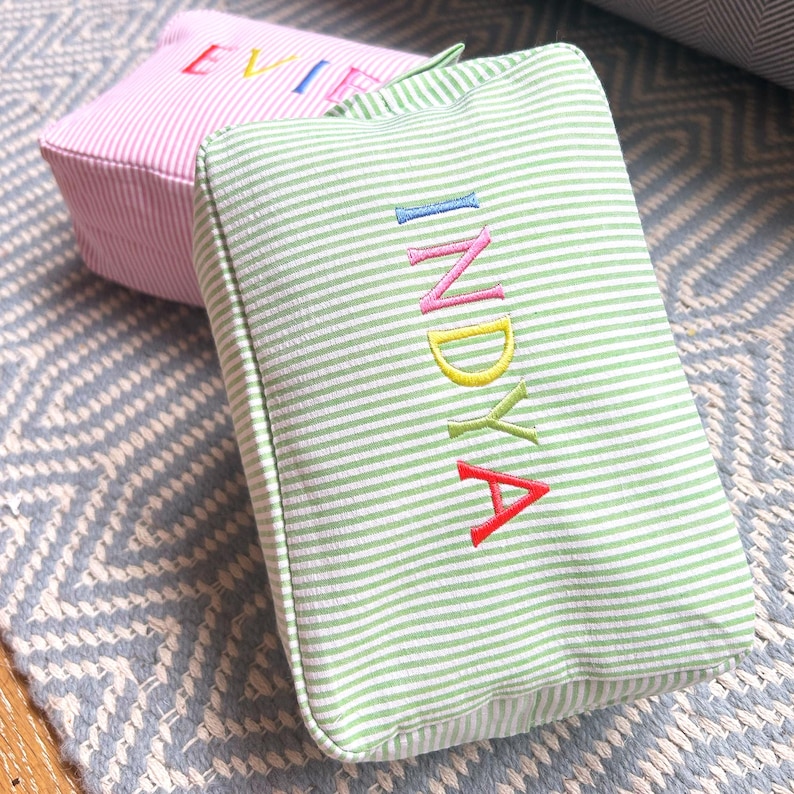 May include: Green and white striped fabric cosmetic bag with the name "INDYA" embroidered in rainbow colors.