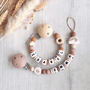 Può includere: Un mordedor per neonati in silicone beige e marrone con un clip e perline che compongono il nome "ISABELLA" e "EMMA". Il mordedor ha anche un ciondolo a forma di girasole e un ciondolo a forma di arcobaleno.
