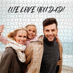 May include: A family of three, a woman, a girl, and a man, are smiling at the camera. The woman is wearing a brown coat and a white scarf. The girl is wearing a pink shirt. The man is wearing a brown coat and a gray sweater. The text "WE LOVE YOU DAD!" is written in black at the top of the image.