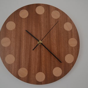May include: A round wooden clock with a brown wood face and light brown wood accents around the edge. The clock has black hands and a gold centre.
