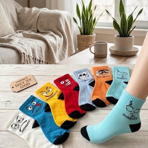 May include: A set of seven ankle socks in white, blue, yellow, red, light blue, orange, and cyan, each featuring a unique cartoon face and the corresponding day of the week printed in black text, with black heels and toes on each sock.