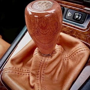 May include: A close-up of a brown leather gear shift boot with a wooden gear shift knob. The knob has a coin embedded in the top.