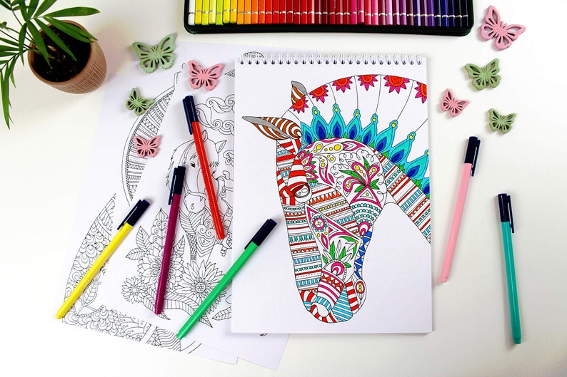 以下が含まれることがあります： 表紙にカラフルな馬の頭を描いた塗り絵。馬は赤、青、緑、黄色の複雑な模様で飾られています。本は空白のページを開いており、塗り絵の準備ができています。