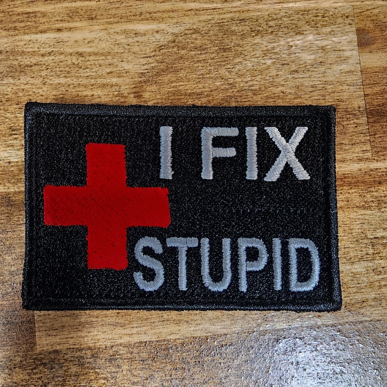 Puede incluir: Parche bordado negro con una cruz roja y el texto "I FIX STUPID".