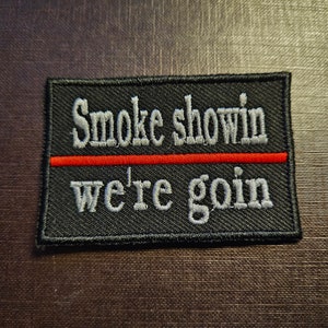 以下が含まれることがあります： 黒い刺繍パッチに白い文字で「Smoke showin' we're goin'」と書いてあり、2行の文字を赤い線が分けている。