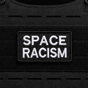 Puede incluir: Un parche rectangular negro con texto bordado en blanco que dice SPACE RACISM. El parche está sujeto a un chaleco táctico negro que presenta correas horizontales y una superficie de tela texturizada con un patrón de rejilla visible.