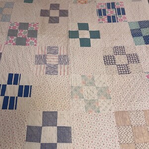 May include: A patchwork quilt with a repeating pattern of cross-shaped blocks. The blocks feature various patterns and colours, including blue, pink, and green, set against a white background with small floral prints. The quilt is made of fabric.