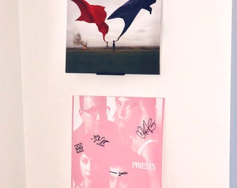 May include: Two framed album covers displayed on a white wall. The top album cover features a red and blue flag design with the text "Biffy Clyro" and "Only Revolutions." The bottom album cover is pink with the band name "Priests" and signatures.