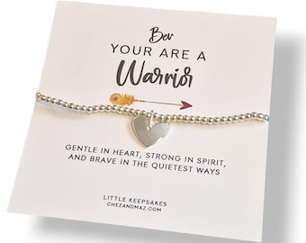 May include: A silver beaded bracelet with a heart charm, displayed on a white card. The card reads "Be Your Are A Warrior" with an arrow graphic and the text "Gentle in heart, strong in spirit, and brave in the quietest ways."
