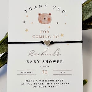 Puede incluir: Tarjeta de recuerdo de baby shower con una pulsera de cordón negro y un dije de corazón plateado. La tarjeta dice "Gracias" y "Baby Shower de Rachael" con la fecha 30 de agosto de 2023. La tarjeta también incluye una linda ilustración de oso.
