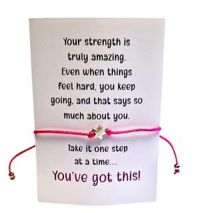 Op de afbeelding: Een roze touw armband met een zilveren ster bedel wordt getoond voor een witte kaart. De kaart heeft de tekst "Your strength is truly amazing... You've got this!"