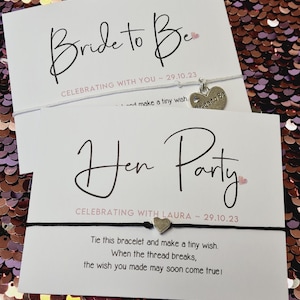 Può includere: Due carte bianche con testo nero e cuori rosa. La carta superiore dice "Bride to Be" e "Celebrating with you ~ 29.10.23". La carta inferiore dice "Hen Party" e "Celebrating with Laura ~ 29.10.23". Entrambe le carte hanno un braccialetto di corda con un ciondolo.