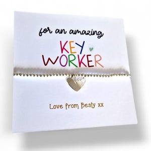 Könnte beinhalten: Ein silbernes Armband mit Herzanhänger liegt vor einer weißen Karte. Auf der Karte steht in bunten Buchstaben "for an amazing KEY WORKER", darunter "Love from Besty xx".