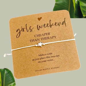May include: A white string bracelet with a silver star charm. The card reads "girls weekend CHEAPER THAN THERAPY" and "Make a wish as you place this bracelet on your wrist... STYLISH SIMPLE ELEGANT".