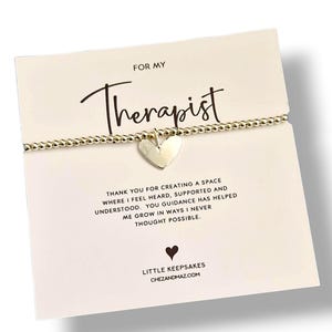 Könnte beinhalten: Ein goldfarbenes Perlenarmband mit Herzanhänger liegt auf einer Karte mit der Aufschrift "FOR MY Therapist". Die Karte enthält eine Dankesbotschaft und die Website chezandmaz.com.