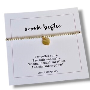 May include: A gold-toned beaded bracelet with a central heart charm, displayed on a white card featuring the text work bestie, For coffee runs, Eye rolls and sighs, Getting through meetings, And sharing supplies, and the brand name LITTLE KEEPSAKES.