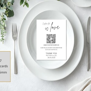 May include: A white rectangular A7 place card measuring 70x95mm with black text and a QR code. The card features the phrase Capture the love, a heart icon, instructions to share photos, and the names Peter & Jo with the date 28.05.2026.
