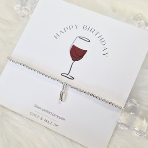 Puede incluir: Un brazalete plateado con un pequeño dije rectangular cuelga de una tarjeta que dice "Happy Birthday" con una ilustración de copa de vino. La tarjeta también dice "Silver plated bracelet" y "CHEZ & MAZ UK". El fondo es blanco con formas decorativas de hielo.