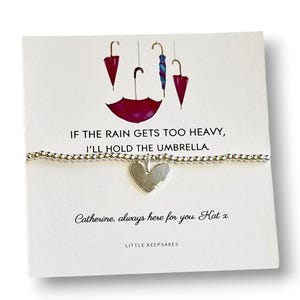 Op de afbeelding: Een zilveren armband met een hartvormige bedel, gepresenteerd op een kaart met paraplu-illustraties. De kaart bevat de tekst "If the rain gets too heavy, I'll hold the umbrella." en een persoonlijke boodschap.