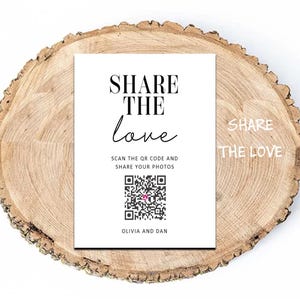 May include: A rectangular white sign with black text that reads SHARE THE LOVE, SCAN THE QR CODE AND SHARE YOUR PHOTOS, and OLIVIA AND DAN, featuring a central QR code with a small pink icon, placed on a circular natural wood slice with bark edges.