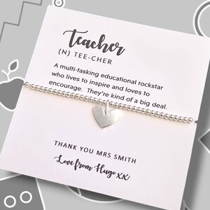 Peut inclure: Un bracelet argenté à perles avec un charm en forme de cœur, présenté sur une carte blanche. La carte affiche le mot "Teacher" avec une définition et un message personnalisé. Le fond est gris.