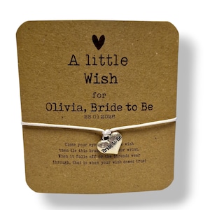 Może przedstawiać: Bransoletka z zawieszką w kształcie serca na brązowej karcie. Na karcie napis "A little Wish for Olivia, Bride to Be" z datą 25.01.2026. Zawieszka mówi "Bride to Be".